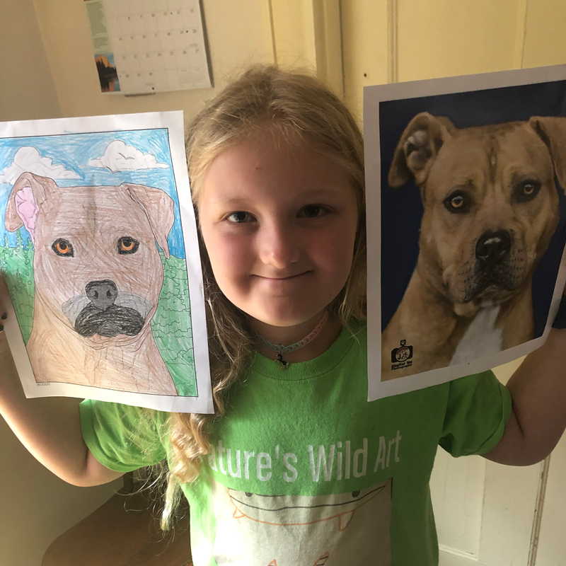 a girl holds her drawing of a dog next to the dog's photo
