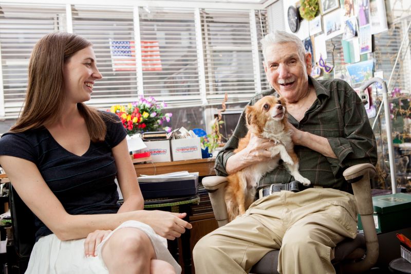 a smiling elderly man holds his dog while a younger woman looks on