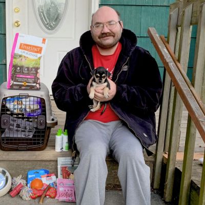 a man holds his puppy while surrounded by pet supplies on his porch
